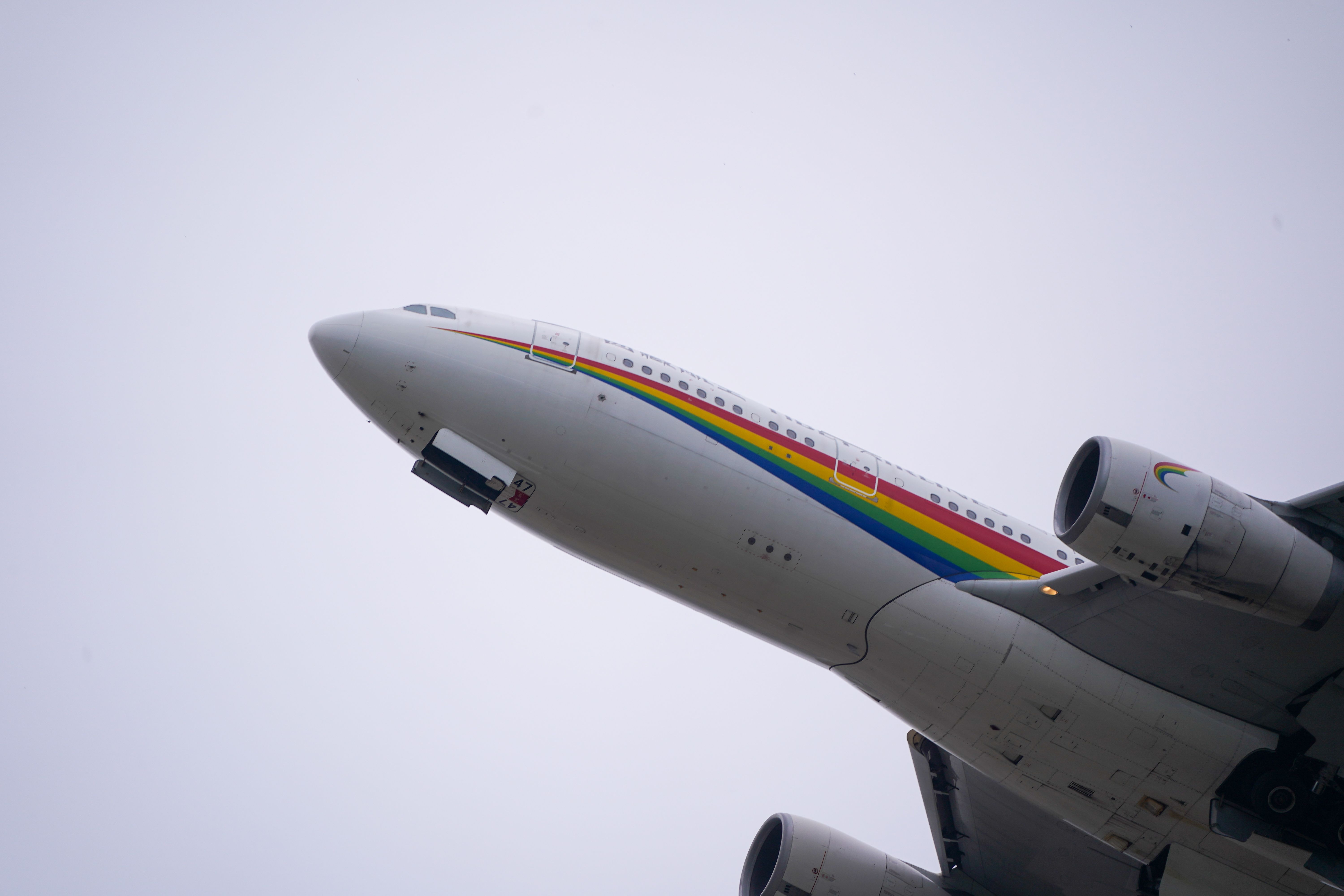 Colorful airplane with a rainbow stripe flies upwards against a cloudy sky.