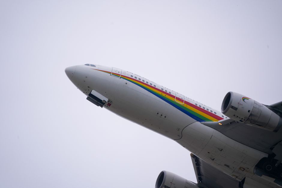 Colorful airplane with a rainbow stripe flies upwards against a cloudy sky.