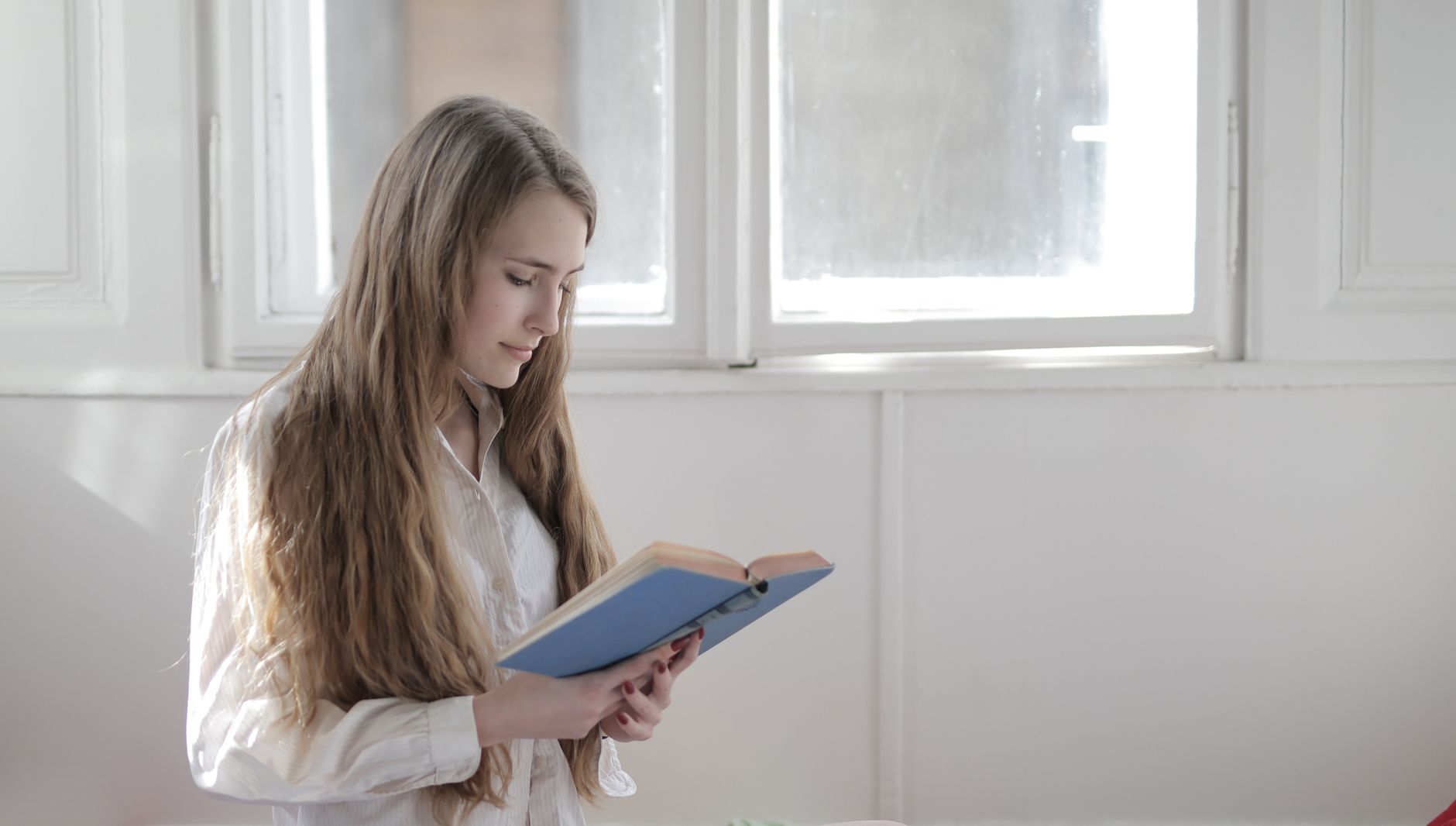 A young woman engrossed in a book while standing near a sunlit window indoors.. pexels, young, woman