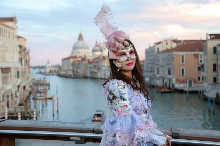 Side view of unrecognizable woman in magnificent costume and gorgeous Venetian mask with feathers standing on bridge in middle of Grand canal against background of cathedral of Santa Maria della Salute in Venice in Italy and looking at camera