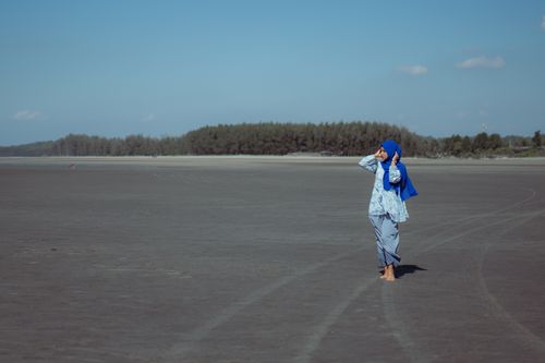 Free A woman in a blue hijab walking on a wide beach with a forested horizon and clear sky. Stock Photo