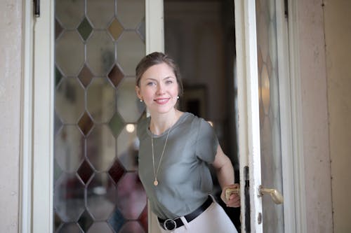 Young smiling female in casual outfit looking at camera and holding on doorknob while entering through stained glass vintage door in ancient building