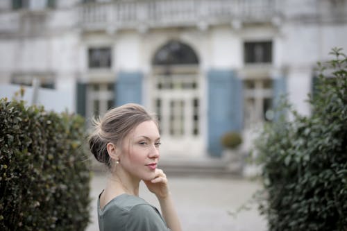 Thoughtful casual lady in gray t shirt with beautiful hairstyle posing and looking away while standing near old building between trimmed bushes of garden