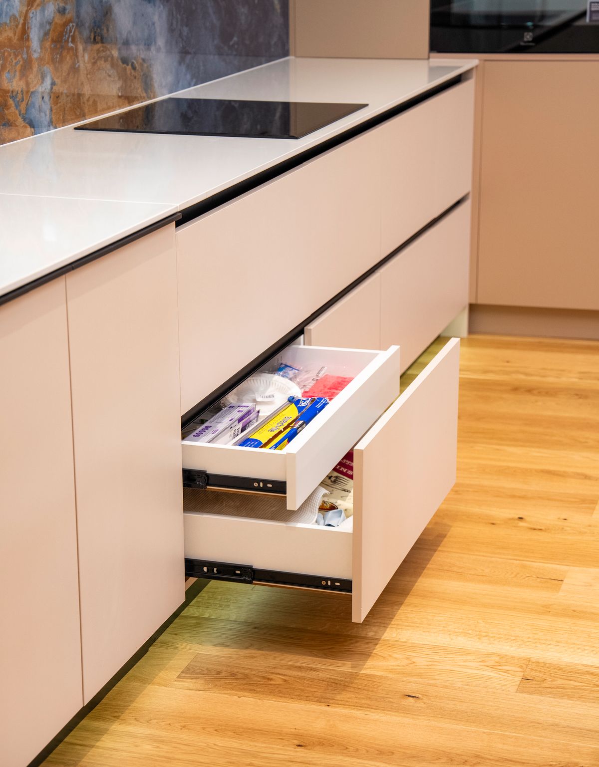 A modern kitchen with pale flat-front cabinetry, a white benchtop, induction cooktop and multiple open drawers over timber flooring.