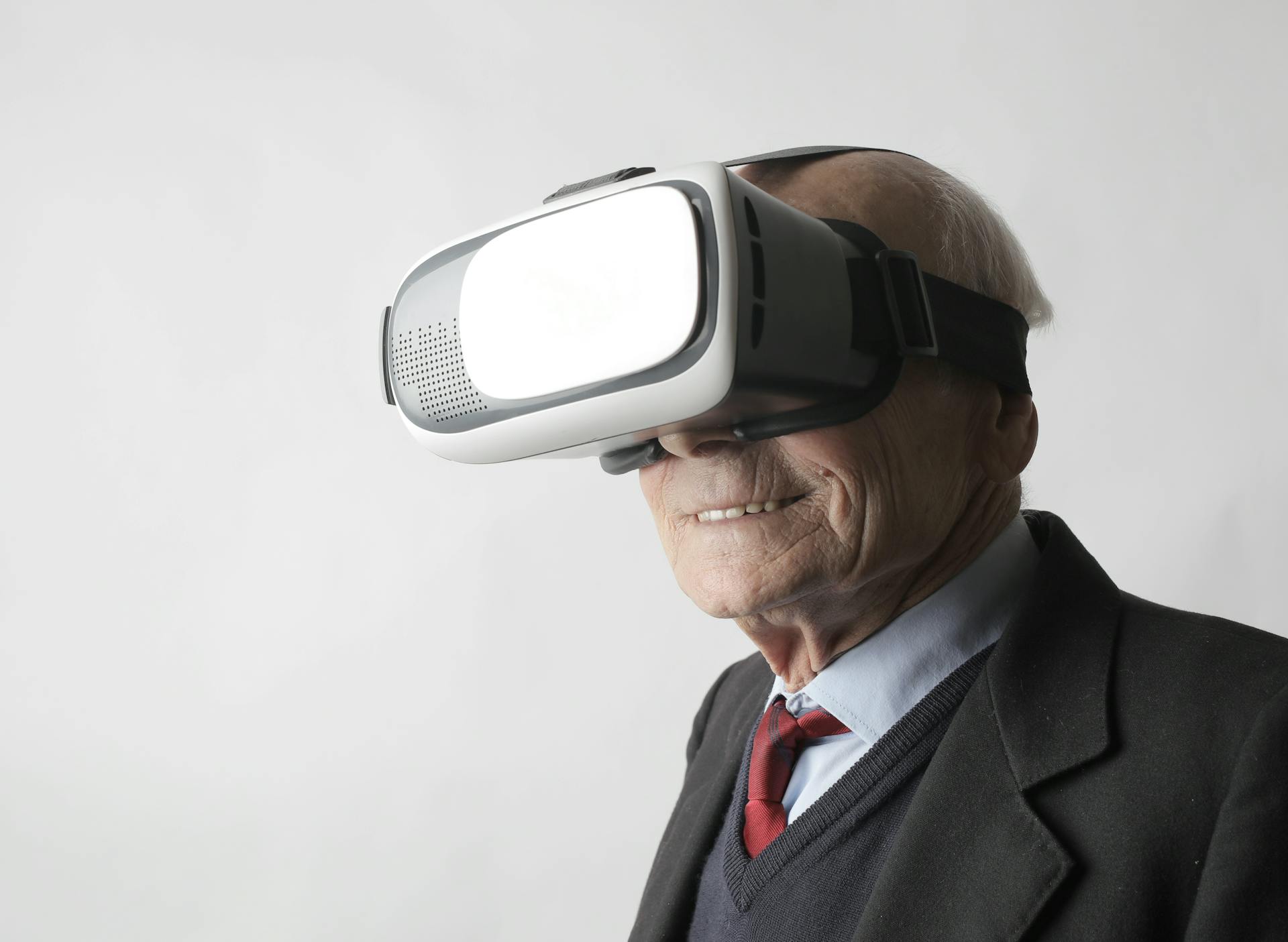 Free Smiling elderly gentleman wearing classy suit experiencing virtual reality while using modern headset on white background Stock Photo