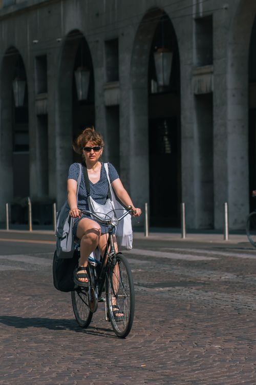 Free A woman rides a bike on a sunlit street with an arched building facade in the background. Stock Photo