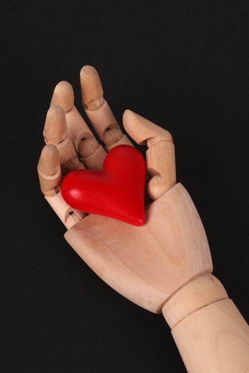 Free A wooden mannequin hand holding a red heart symbol against a black background. Stock Photo