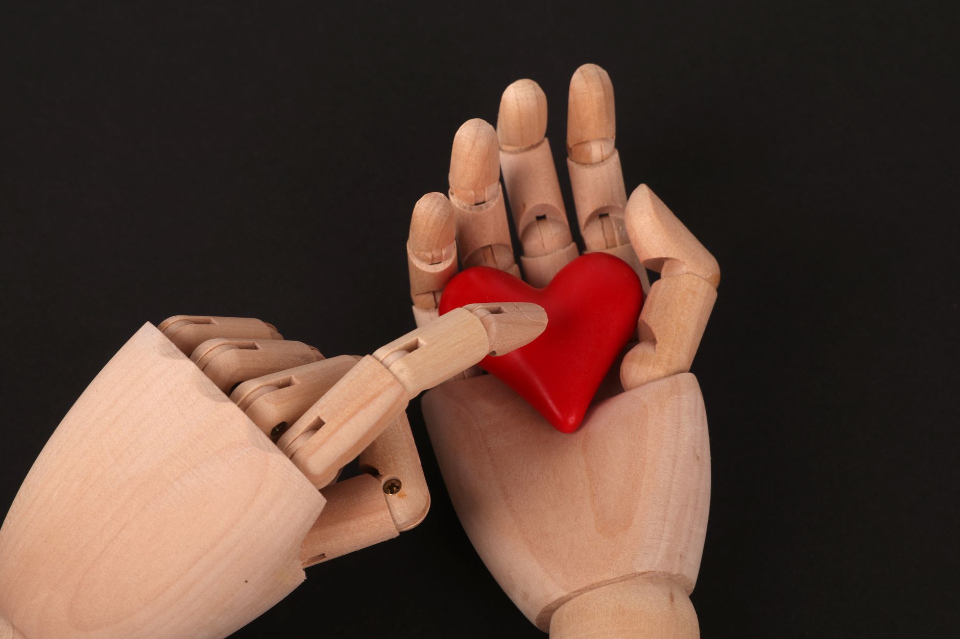 Wooden mannequin hands gently hold a small red heart on a black background, symbolizing care and affection.