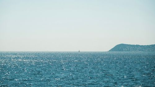 Free A serene view of the open sea with a distant sailboat and a hilly horizon under a clear sky. Stock Photo