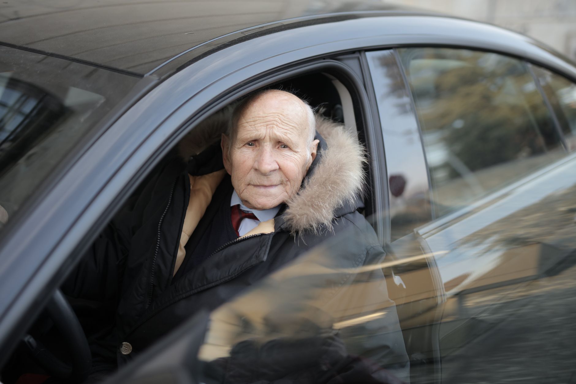 Senior man driving a car, wearing a winter coat, captured through the vehicle window.