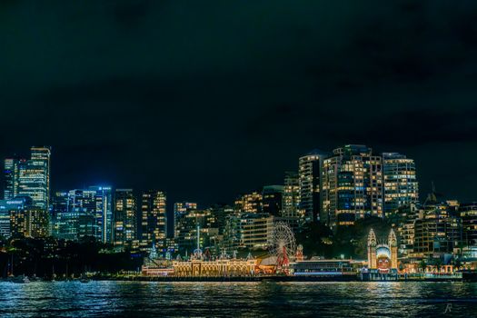 Vivid night view of Sydney's Luna Park with illuminated skyline reflecting in harbor waters, showcasing urban vibrancy.