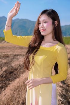 Portrait of a woman in a yellow Ao Dai waving in a sunny field with mountains.