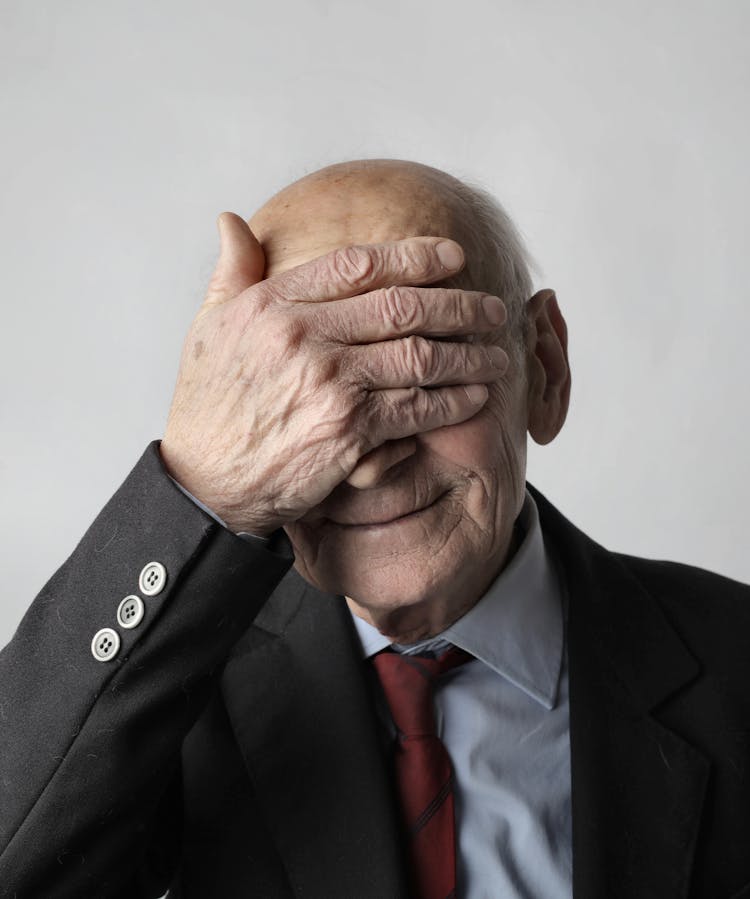 Elderly Man In Black Suit Jacket Covering His Eyes With His Hand