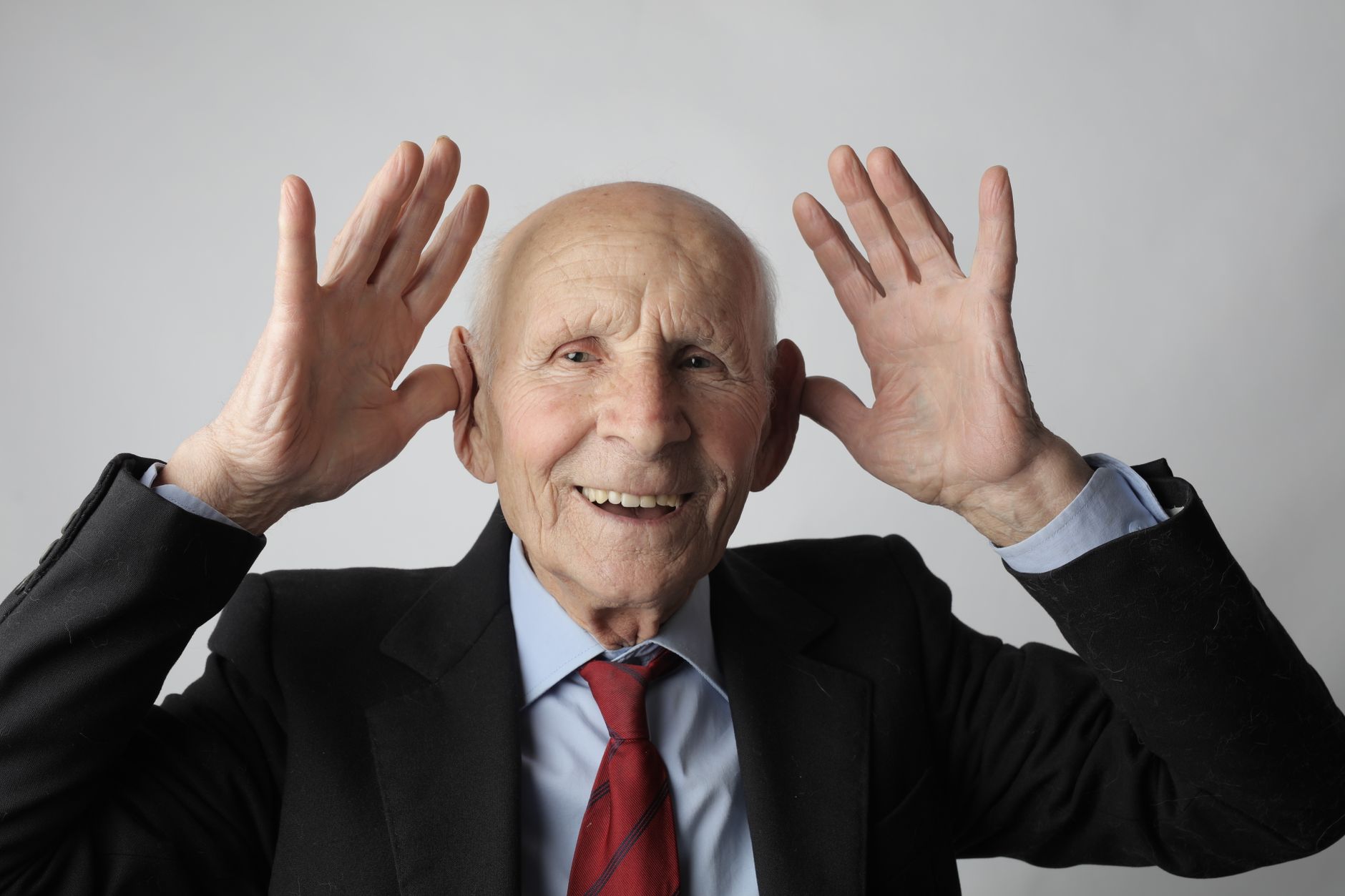 A cheerful elderly man in a suit striking a playful pose with his hands raised beside his head.