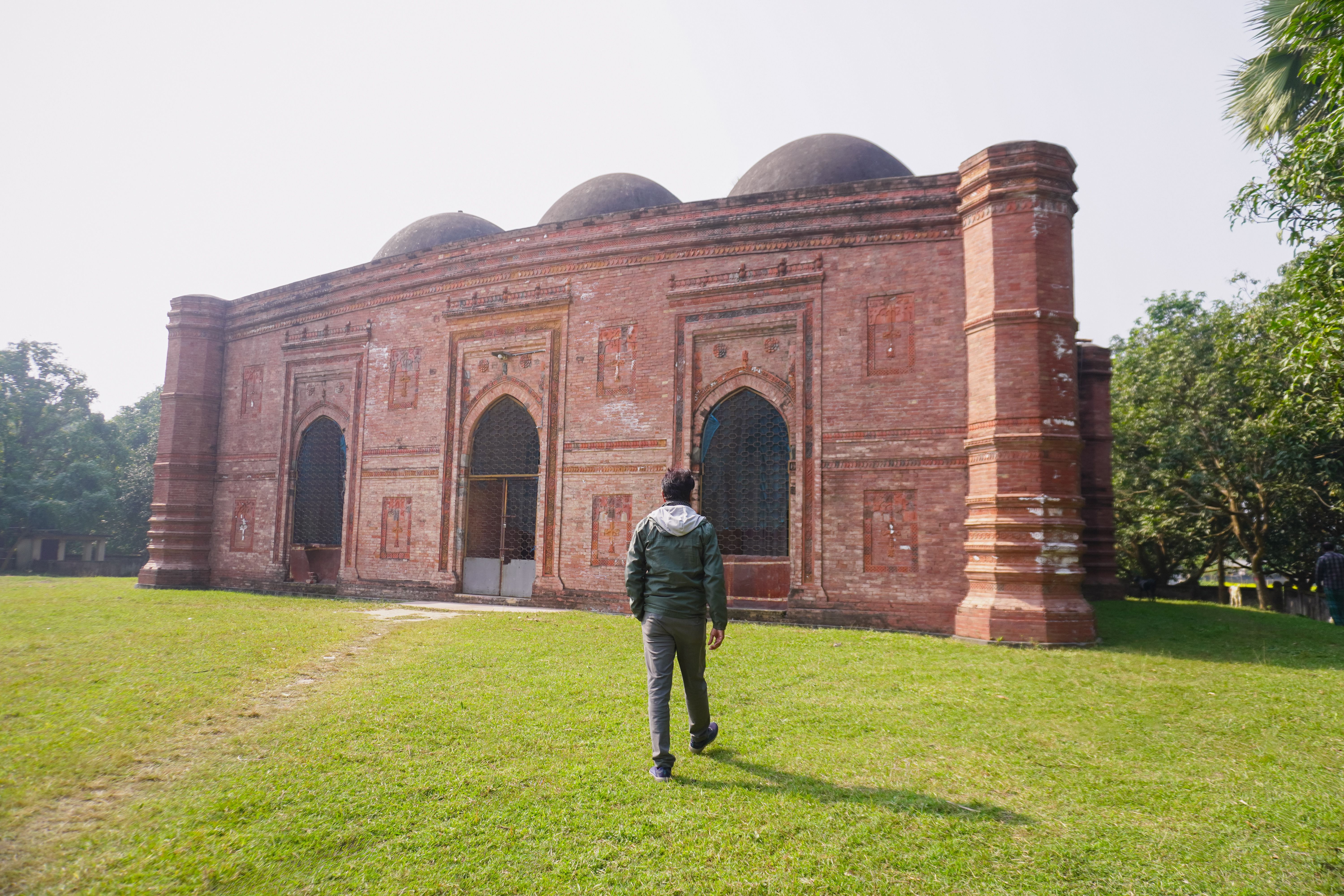A person walking towards a historic red brick mosque with domes on a sunny day.