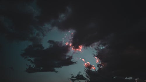 From below of black fluffy clouds with pink gleams of setting sun in dark sky