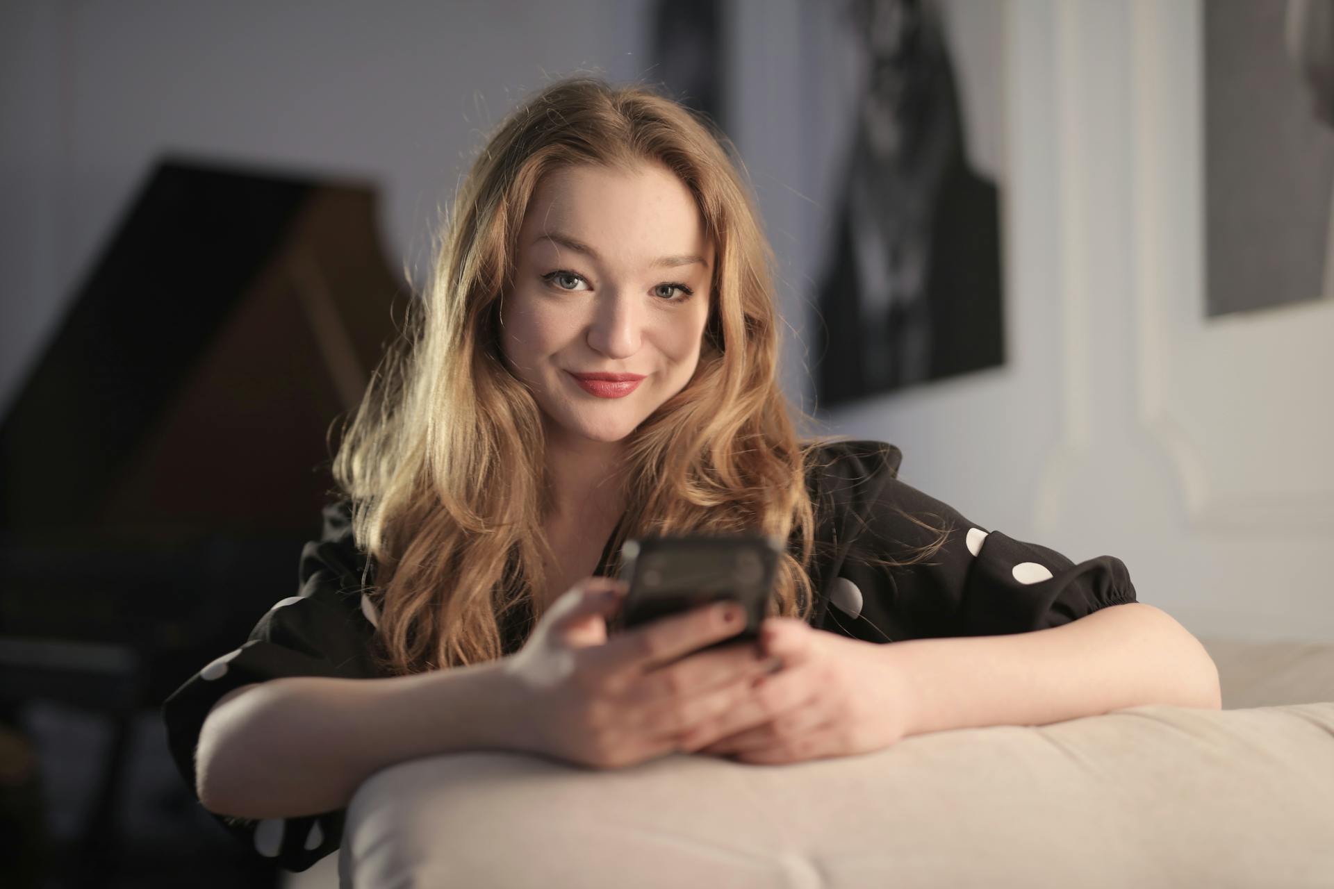 Content female wearing polka dot clothes sitting on sofa and chatting on social media via mobile phone while chilling and looking at camera