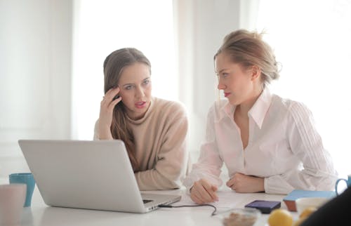Free Pensive female entrepreneurs wearing casual clothes sitting together at table with laptop and talking about problems in project Stock Photo