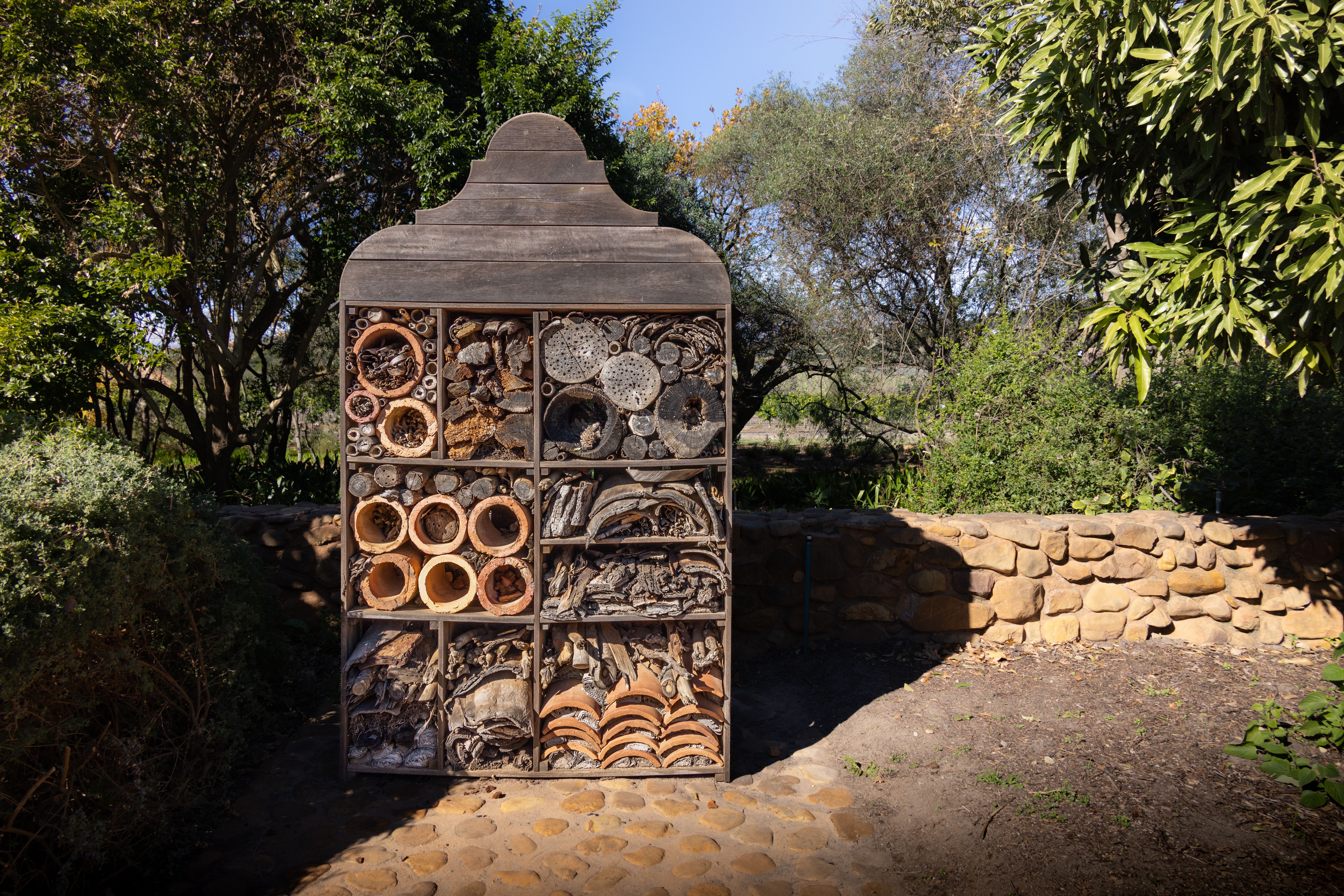 A wooden insect hotel in a garden setting, surrounded by lush greenery, under a bright day.