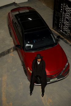 Stylish individual in elegant fashion poses with a luxury red car in an urban setting.