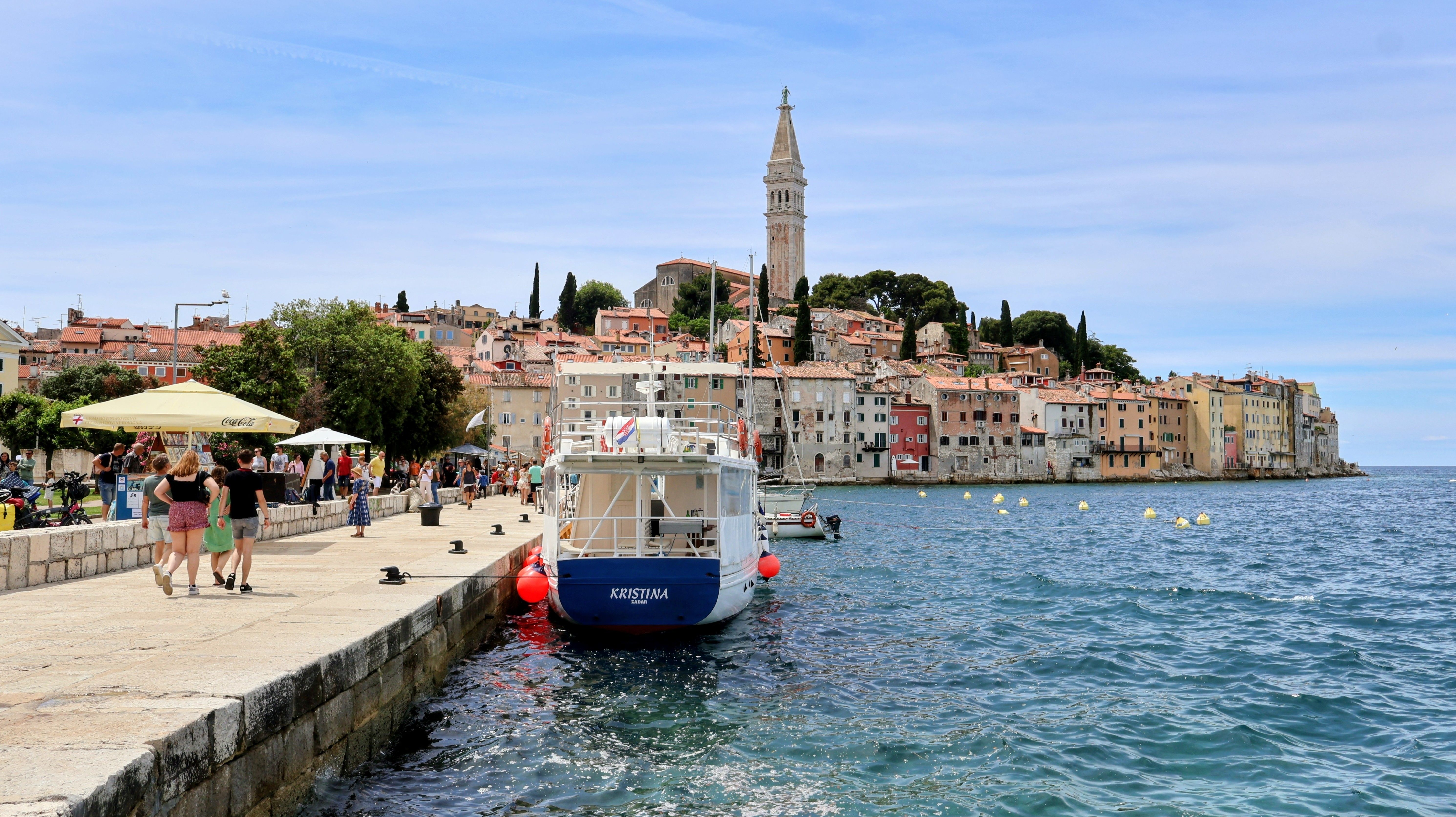 Charming scene of Rovinj harbor showing a docked boat, tourists, and historic bell tower in summer.