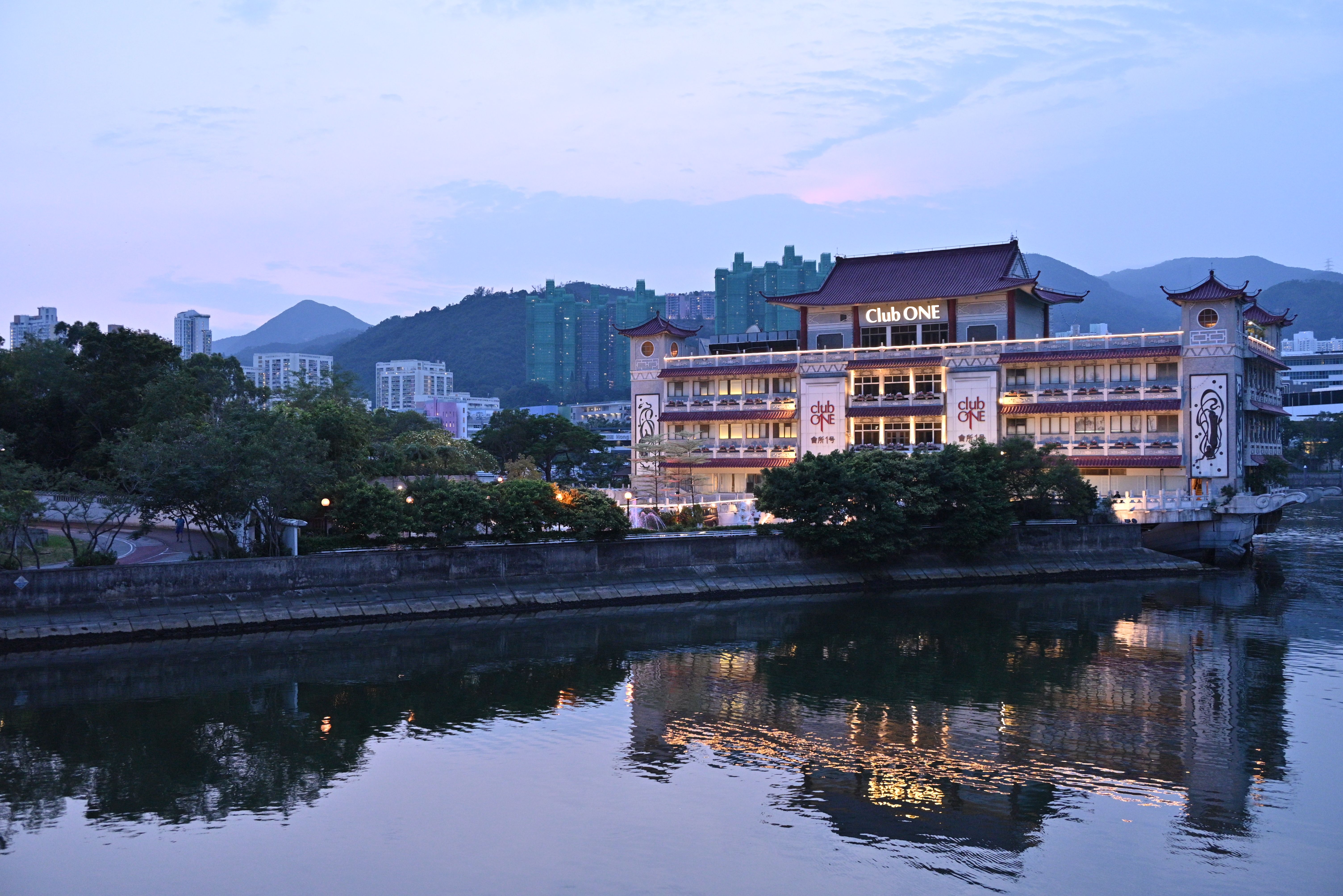 Club ONE building reflected on calm lake at twilight under clear sky, surrounded by mountains.