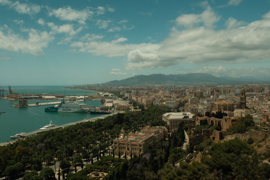 Una vista panorámica del puerto y la ciudad de Málaga con un telón de fondo montañoso bajo un cielo parcialmente nublado.
