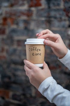 特写镜头中，一只手拿着咖啡杯，背景是砖墙，上面有“咖啡时间”的文字
