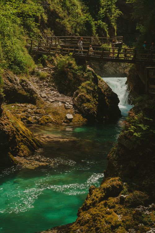 Free Picturesque view of a wooden bridge spanning a vibrant turquoise mountain stream, surrounded by lush greenery. Stock Photo