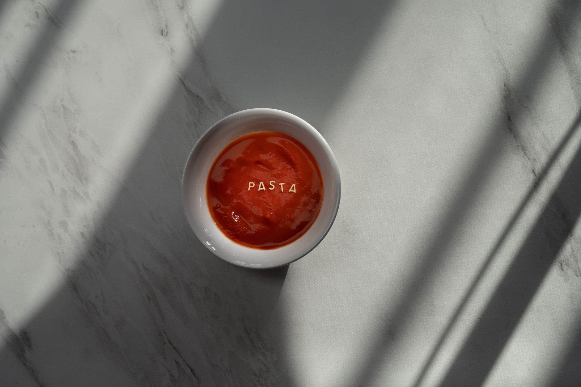 A minimalist close-up of tomato soup with pasta letters on a marble background, warm light.