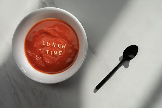 平躺在大理石表面的番茄汤图像中拼出了“午餐时间”字样，并配有一把黑色勺子