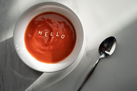 Tomato soup in a bowl with the word 'hello' formed by pasta letters, alongside a spoon.