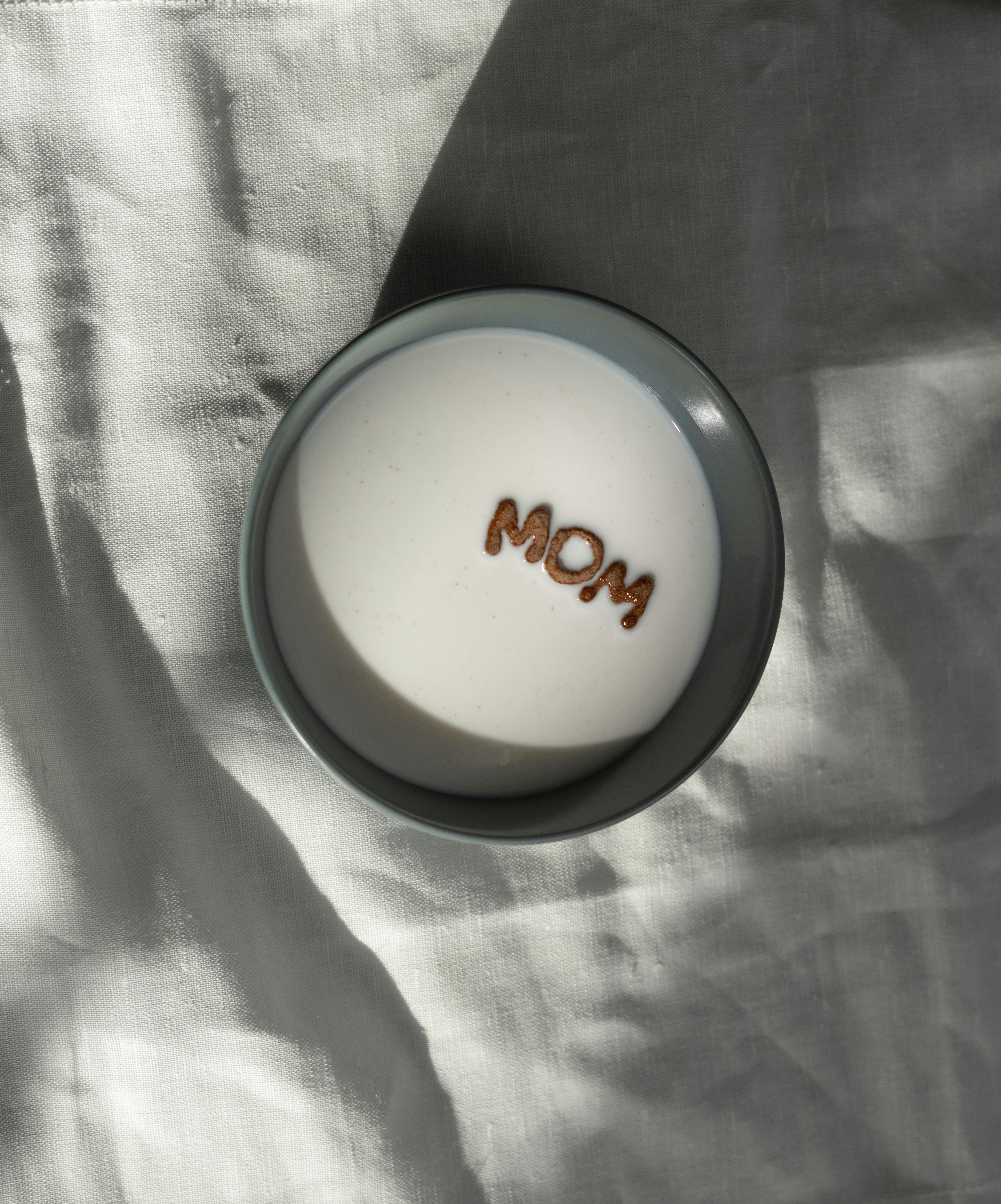 白色织物背景上，一碗牛奶与谷物拼出“妈妈”字样，顶视图