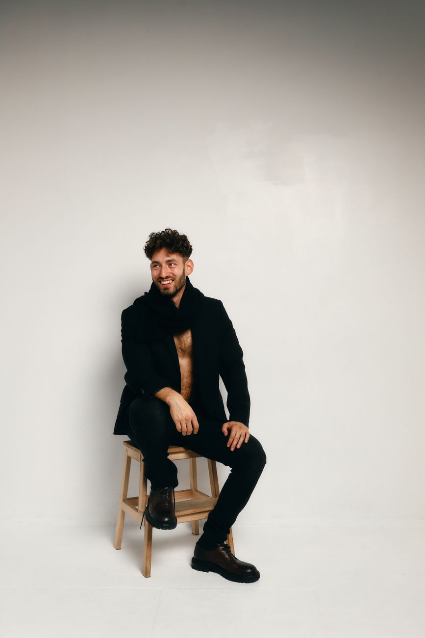 Confident male model in a black outfit and boots poses against a white backdrop, exuding modern style.