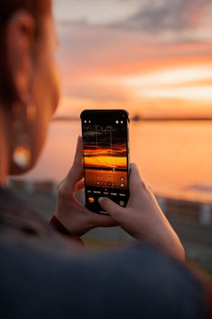 Person photographing a vibrant sunset over water using a mobile phone.