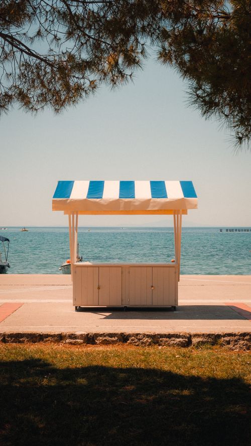 Free An empty vendor stand by the seaside with a blue and white striped canopy under a clear sky. Stock Photo