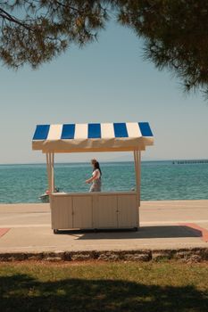 A woman stands at a seaside kiosk on a sunny day with clear blue waters in the background.