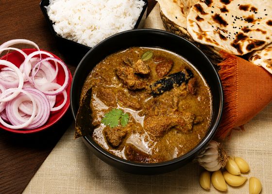 Black bowl of mutton curry with bay leaf and cilantro, naan, rice and red onion