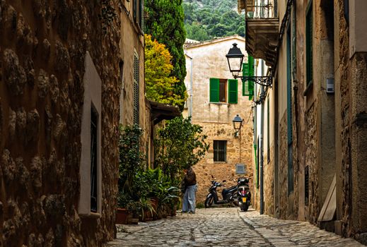 Quaint cobblestone street with rustic walls, greenery, and parked scooters in a picturesque European village.