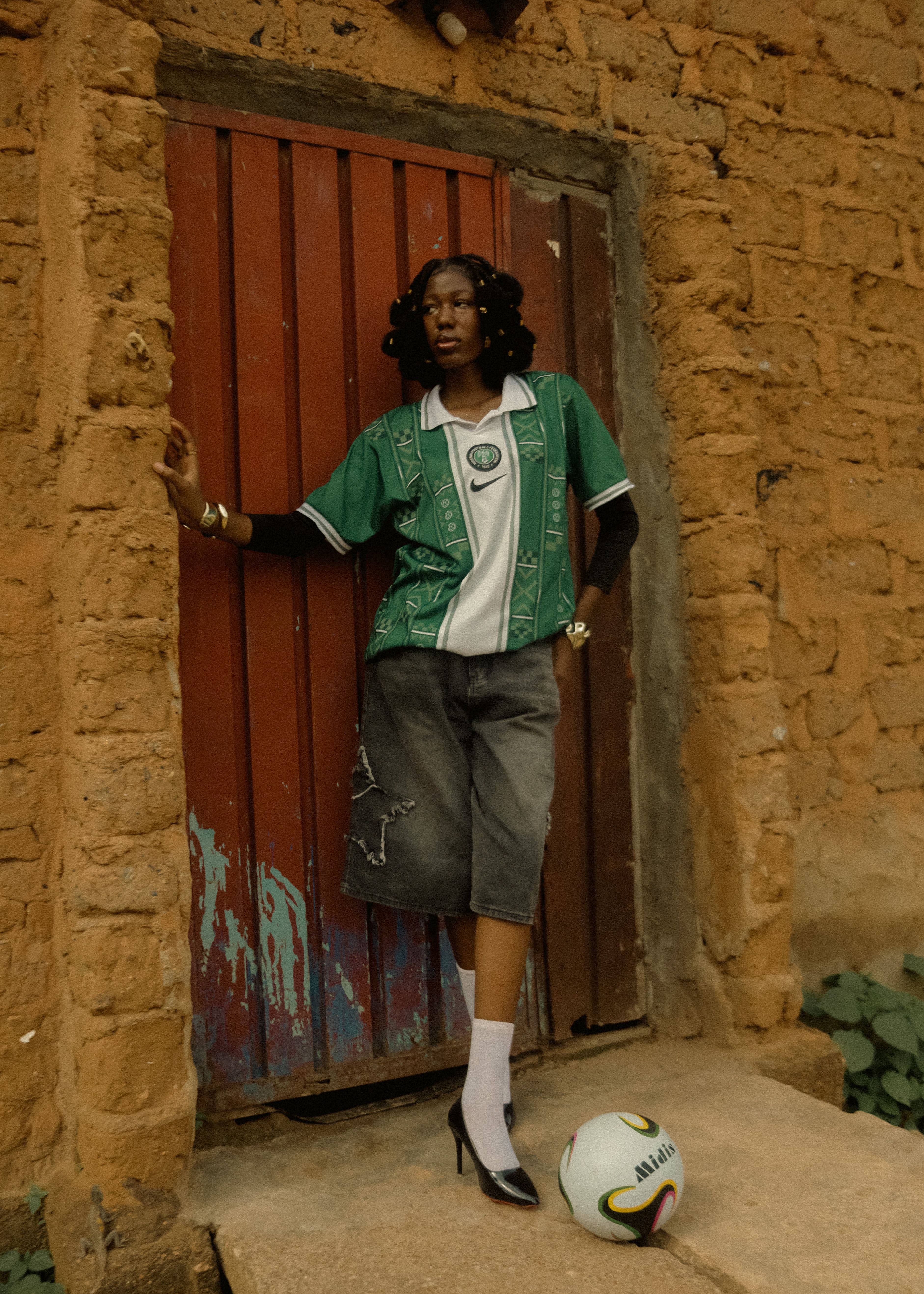 A fashionable adult poses in urban clothing and soccer attire, leaning against a rustic wall.