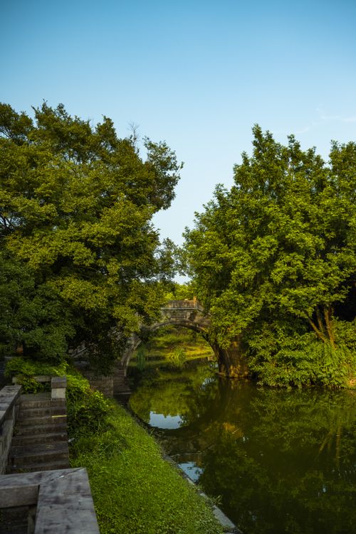Free A serene stone bridge arches over a peaceful river surrounded by lush trees. Stock Photo