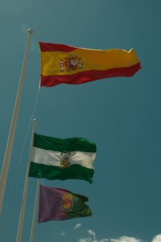 Waving flags of Spain, Andalucía, and Córdoba against a clear blue sky on a sunny day.