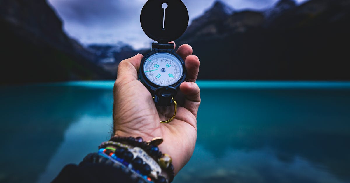 Photo by Joshua Woroniecki A hand holding a compass with a serene lake and mountains in the background, capturing the spirit of adventure.