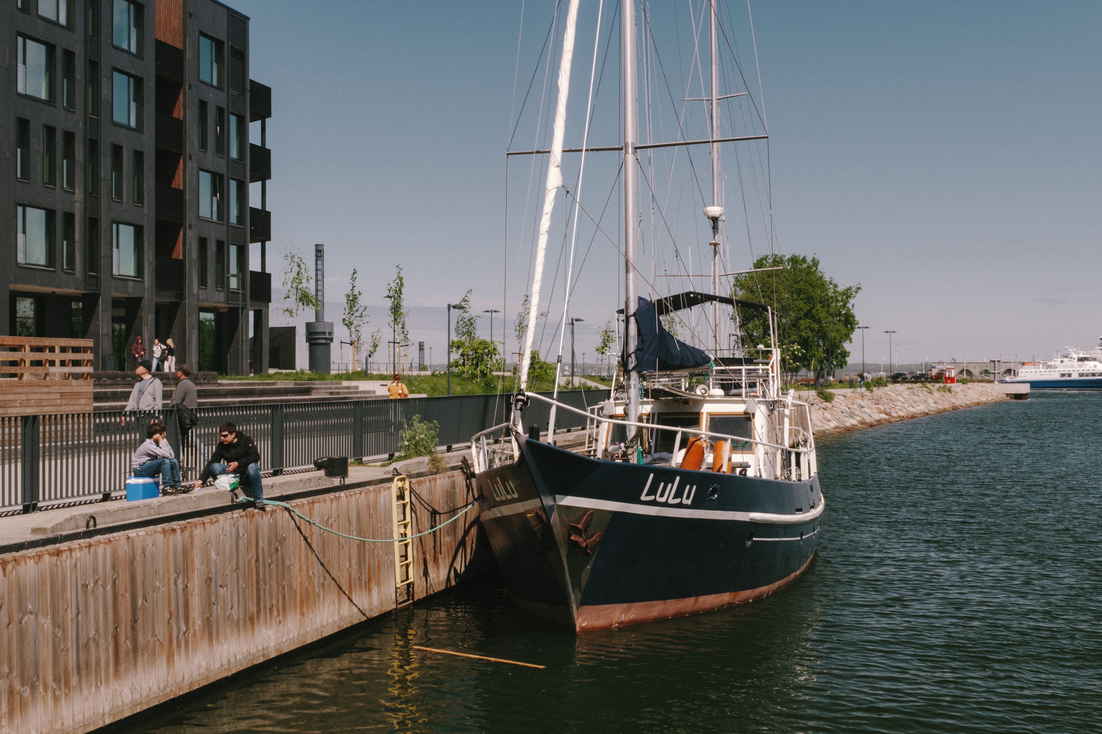 A sailboat named Lulu docked in Tallinn's harbor, with modern buildings and people nearby on a sunny day.