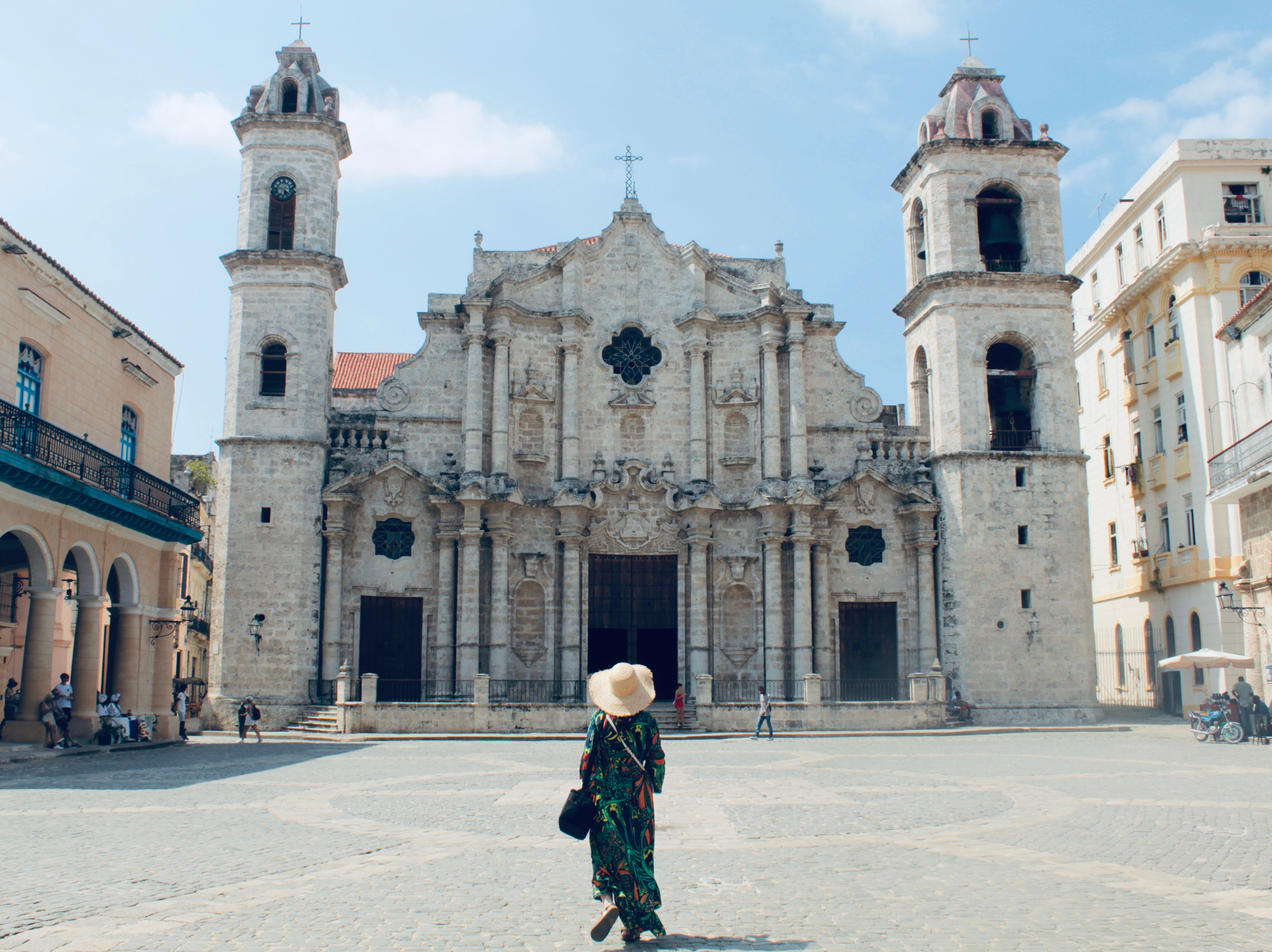 A woman in a sun hat stands before a historic cathedral in Havana, Cuba, admiring its Baroque architecture.