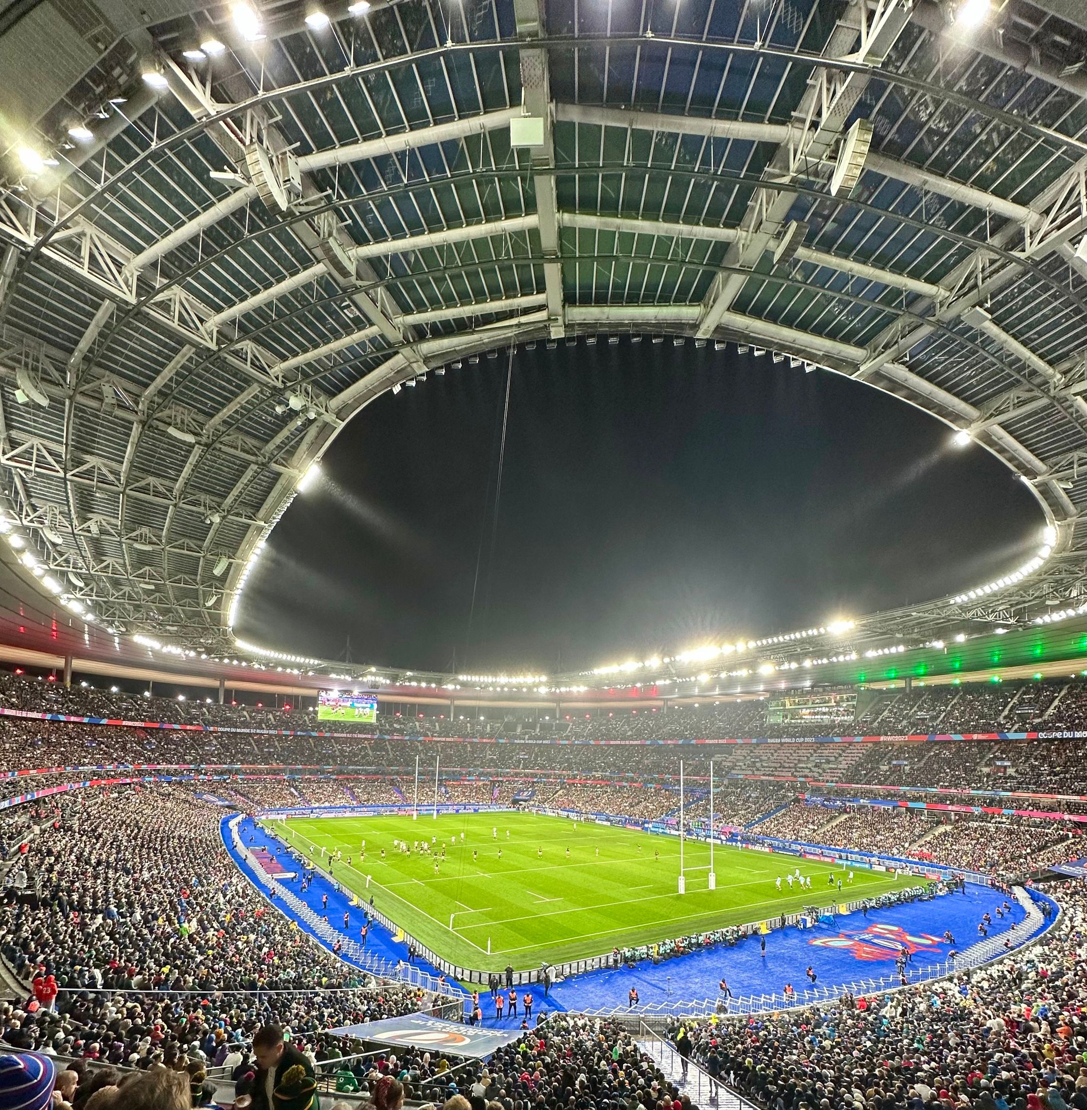 Nighttime panoramic view of a packed rugby match at Stade de France with dazzling lights and vibrant atmosphere.