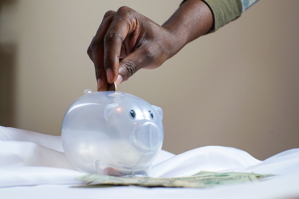 Mão colocando moedas dentro de um cofrinho transparente, representando o hábito de guardar dinheiro todo mês