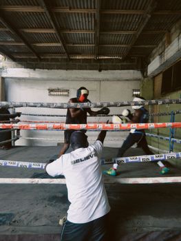 Two boxers sparring in an indoor training ring in Ghana, observed by a coach.