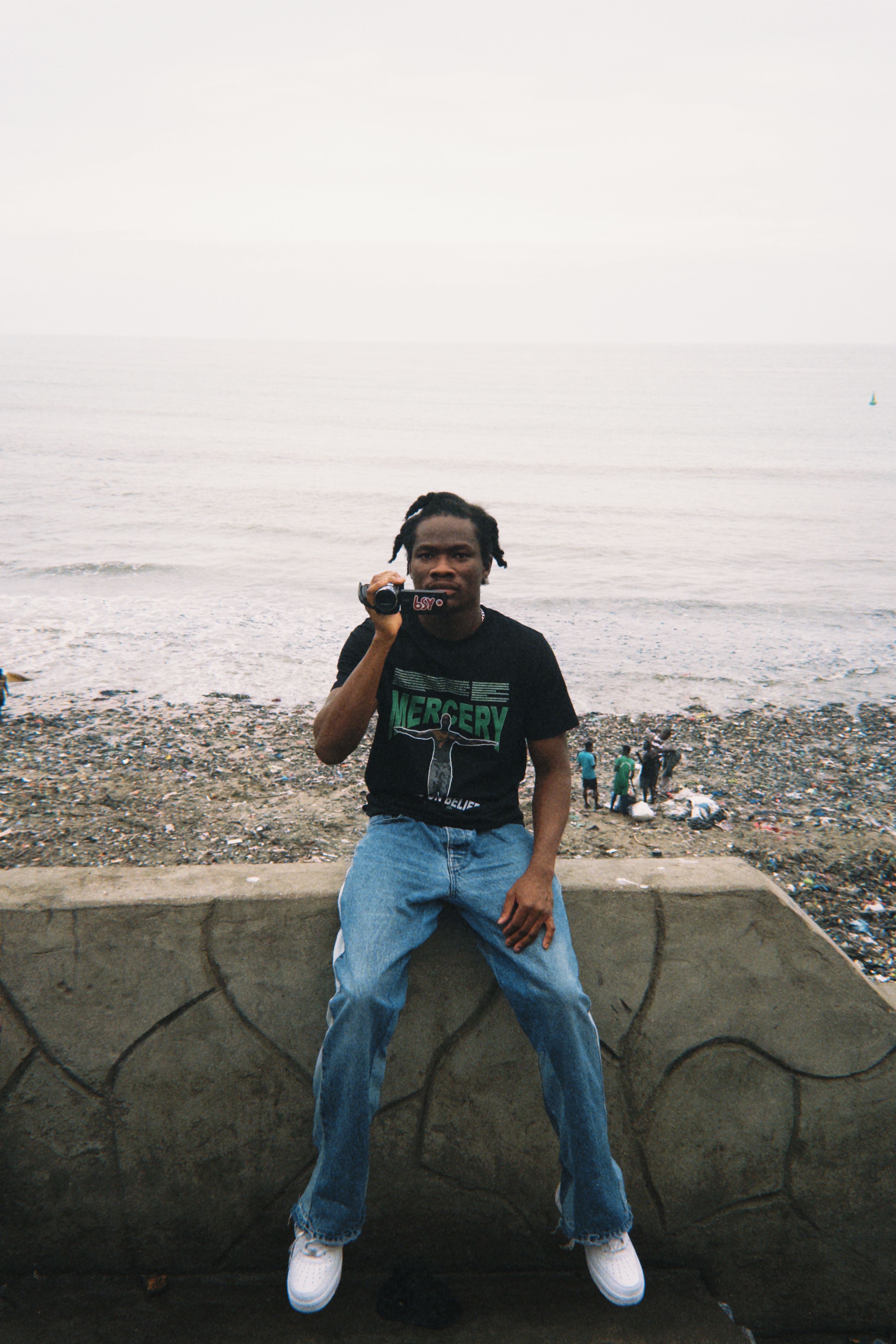 A young man sits on a wall overlooking the beach and ocean on a cloudy day in Ghana.
