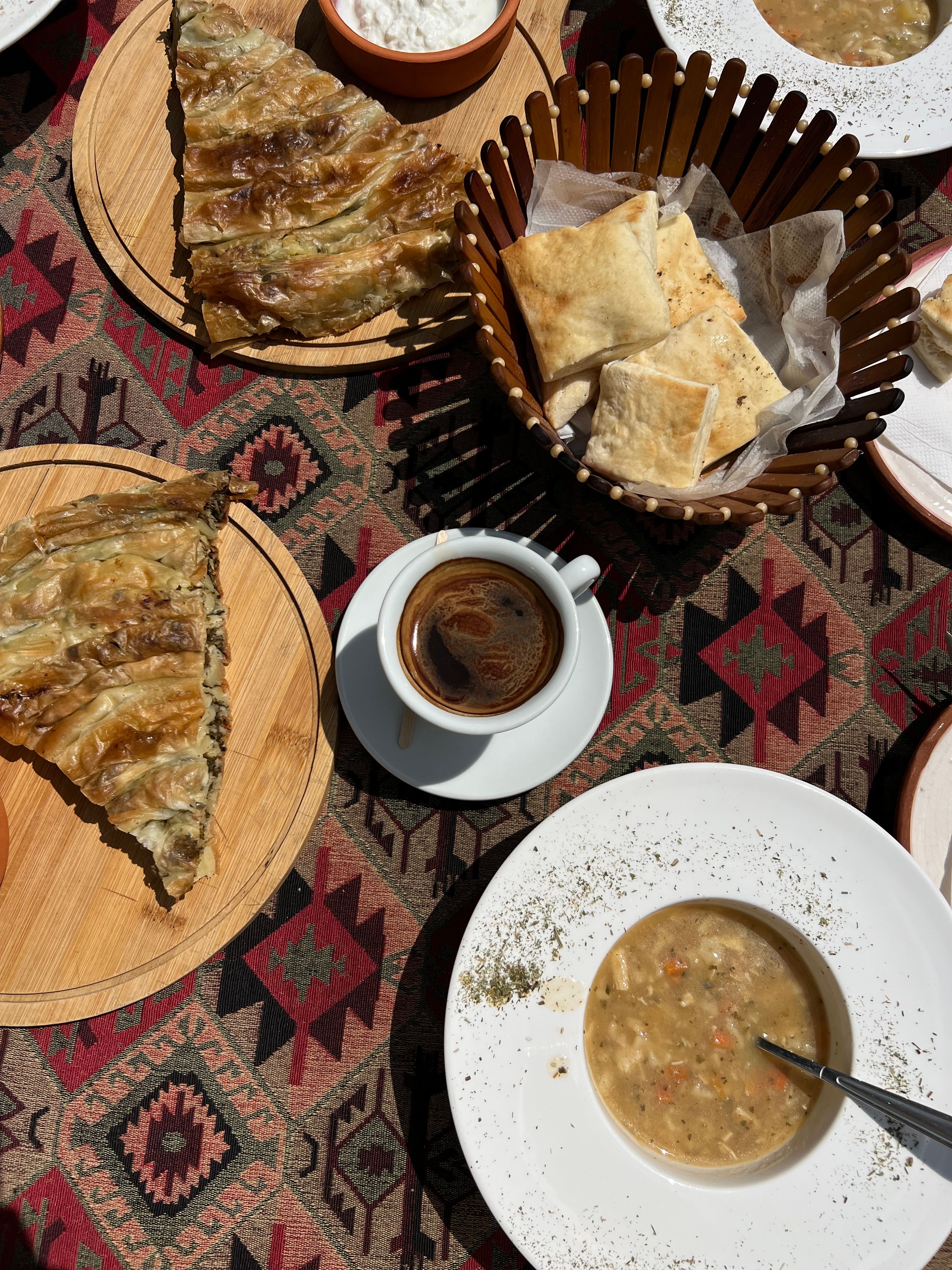 A delicious traditional Montenegrin meal including bread, pastries, soup, and coffee, served on a patterned tablecloth outdoors.